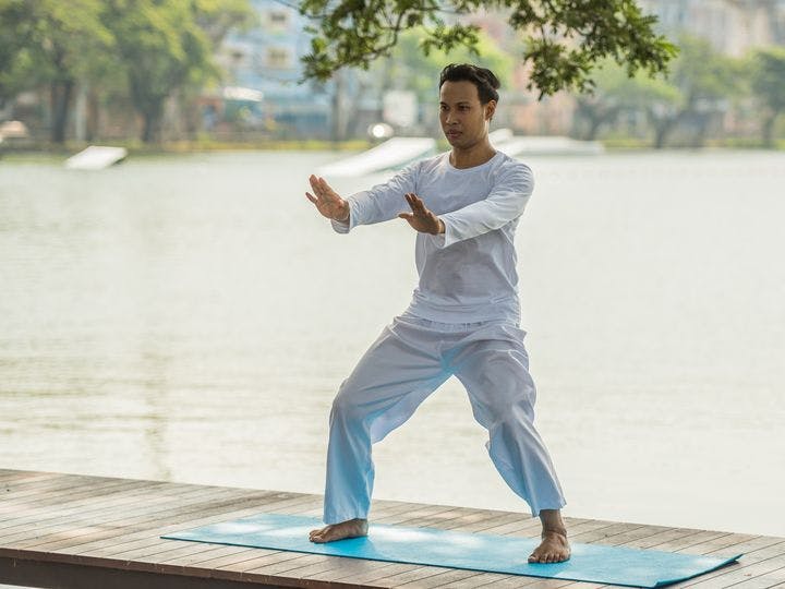 Man wearing white long-sleeved shirt and pants practices Qigong on a yoga mat next to a lake.