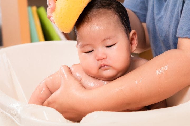 Woman uses a yellow-coloured sponge to bathe an infant in a bathtub, supporting his body with her left hand.