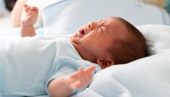 Infant crying while lying on a bed with his arms outstretched.