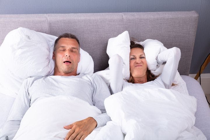 Woman lying in bed covering her ears with a pillow while man next to her snores.