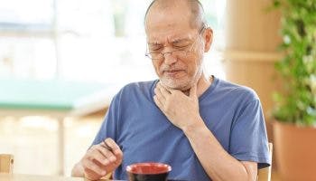 A man in old age holds his throat, having difficulty swallowing while eating.