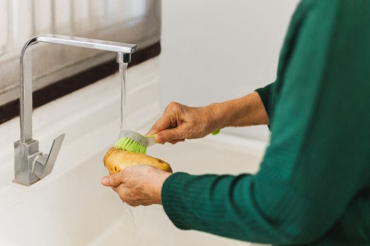 A woman scrubbing a potato with a brush under running tap water.