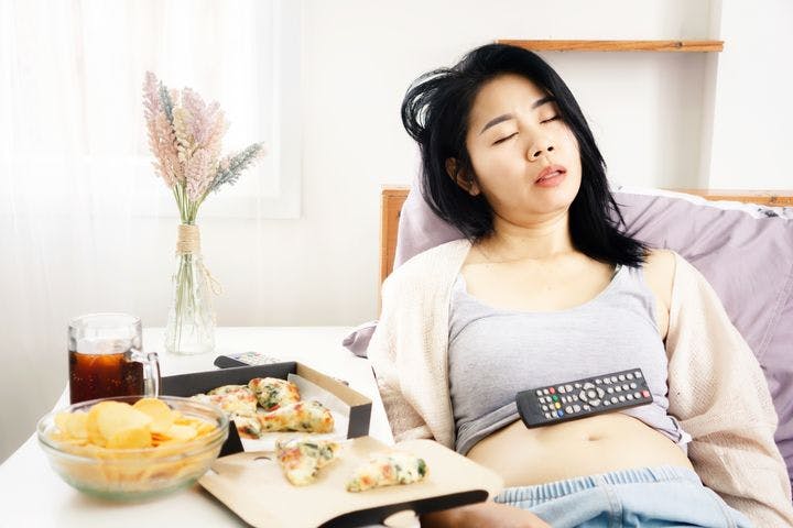 Woman falls asleep with the TV remote on her belly, next to a table filled with pizza, chips, and a soft drink.