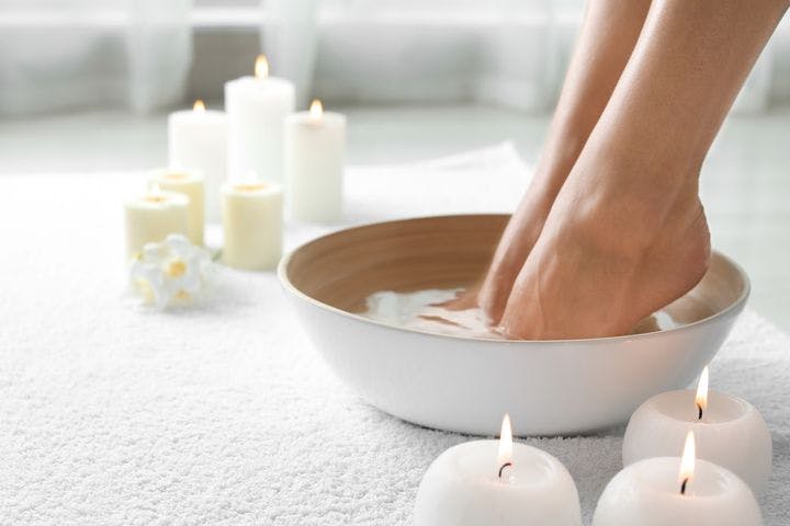 A woman soaking her feet in a bowl of warm water set on a white towel surrounded by candles.