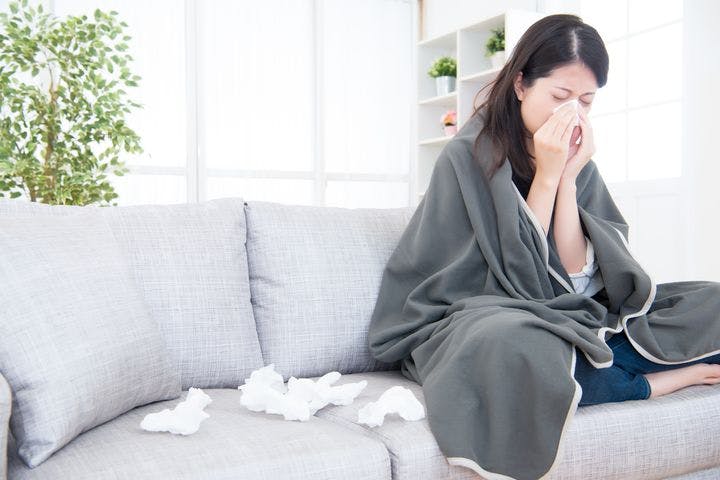 A woman wrapped in a blanket wiping her nose with a tissue while sitting on a sofa next to used tissues.