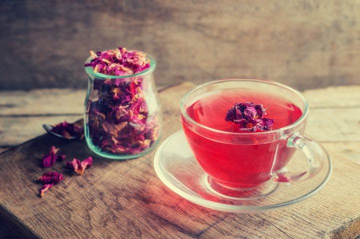 Cup of rose tea next to a jar of dried rose petals on a rustic wooden table.