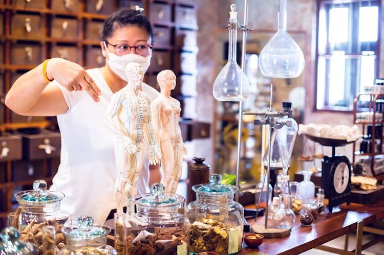 Chinese medicine physician stands behind a counter with herbs, lab equipment, and acupuncture models.