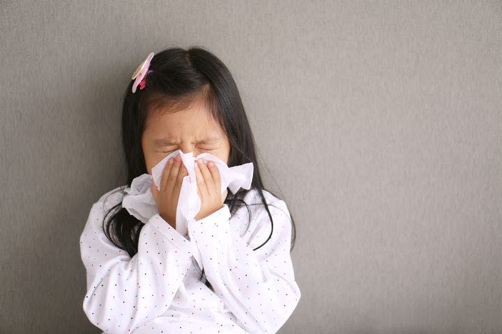 A girl sneezing into a tissue that she’s holding up to her nose with both hands. 