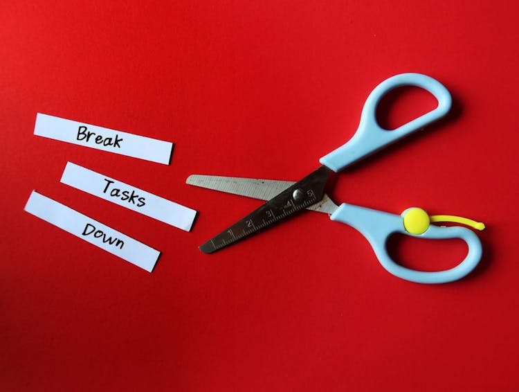 Three pieces of paper with the words “break”, “tasks”, and “down” handwritten on them next to a pair of scissors against a red background.
