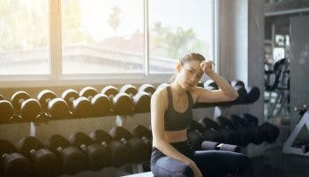 Woman wiping sweat from her forehead sits on a bench in a gym next to weights.