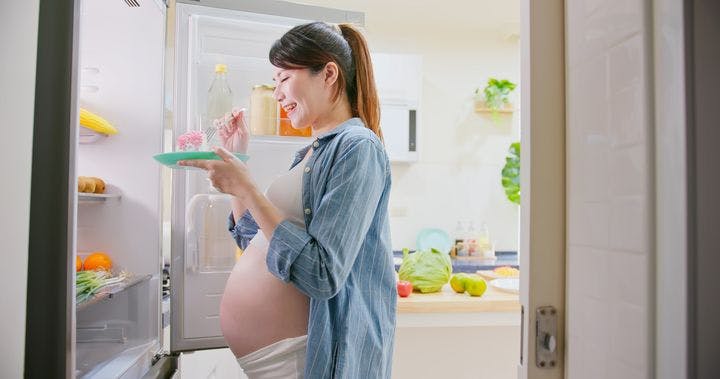 Pregnant woman standing in front of open refrigerator, eating cake