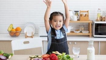 Toddler girl raises her arms and smiles standing in the kitchen in front of a cutting board filled with healthy colourful vegetables and fruits.