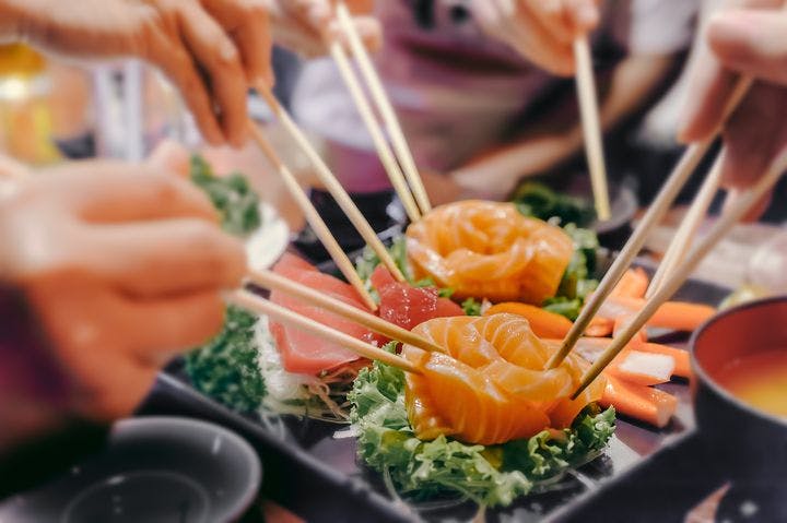 Hands holding chopsticks to pick pieces of salmon sashimi off a tray.