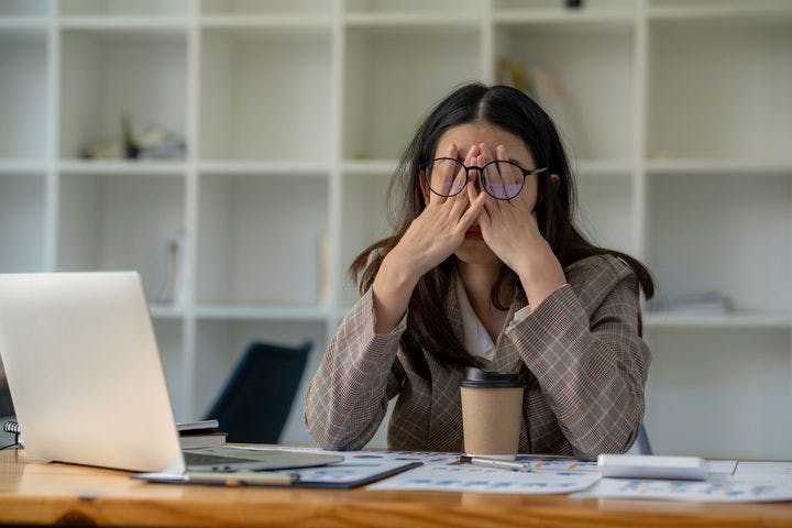A woman covering her eyes with both hands while wearing a pair of eyeglasses as she sits a desk. 