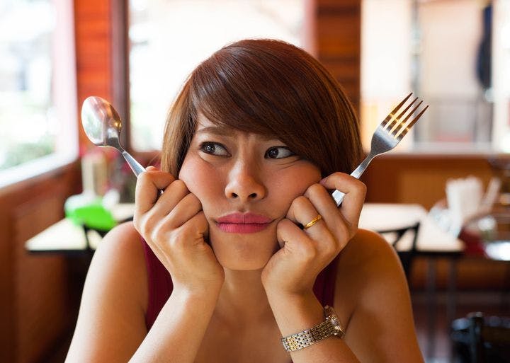 Woman frowns with a confused expression holding a fork in one hand and spoon in the other hand close to her face.