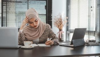 Woman sitting in office writing in her notebook and holding her head in concentration.