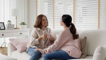 Two middle-aged Asian women face each other to talk while sitting at a sofa.