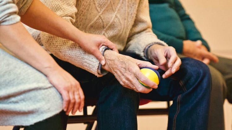 A caregiver's hand holding an elderly's hand, who is holding on to a stress ball.
