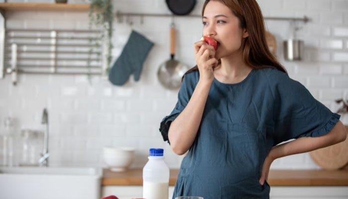Pregnant woman eats an apple while standing in the kitchen.