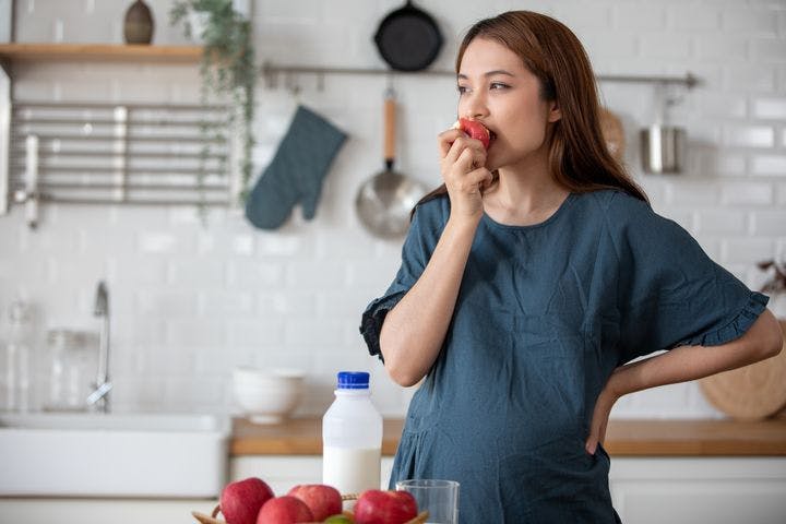 Pregnant woman eats an apple while standing in the kitchen.