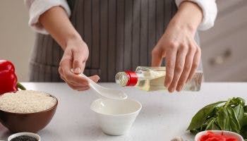 A partial view of a woman’s hands holding a bottle of rice vinegar, preparing to pour it on a spoon and a bowl underneath it.