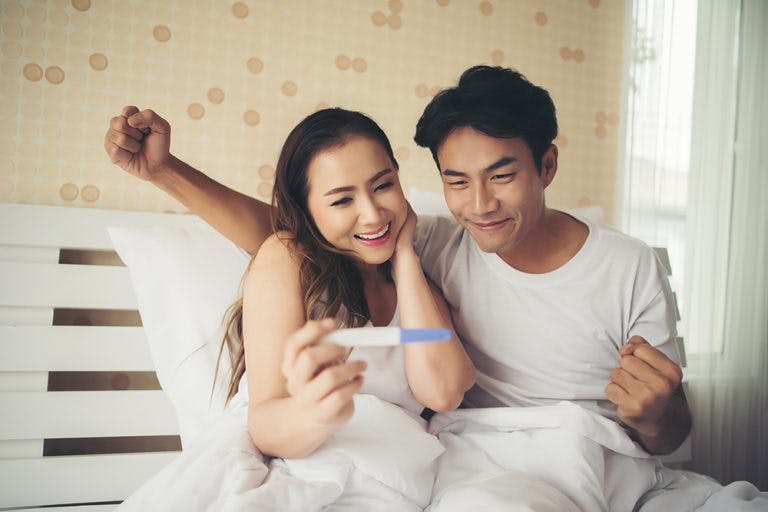 An Asian woman and an Asian man react happily to the result of a home pregnancy test kit while sitting in bed