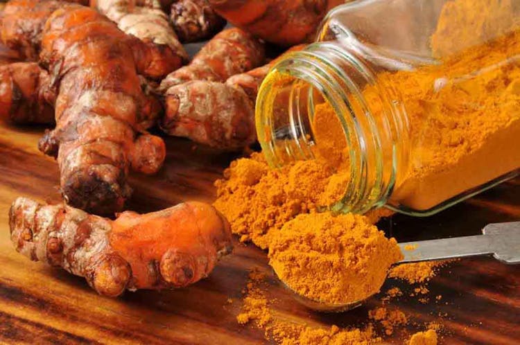 Turmeric roots and powder displayed on a wooden table alongside an open glass container and metal spoon