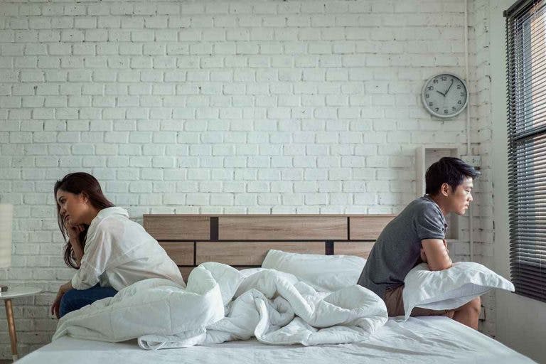 An Asian man and an Asian woman sit on opposite sides of a bed with their backs facing each other