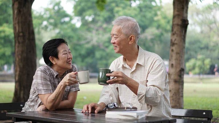 Two senior citizens sit at a picnic table in a park, having a drink together