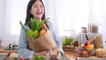 An Asian woman laughs and holds fruits and vegetables in the kitchen