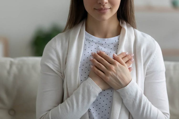 A woman sits down on a sofa and touches the heart area of her chest with both hands