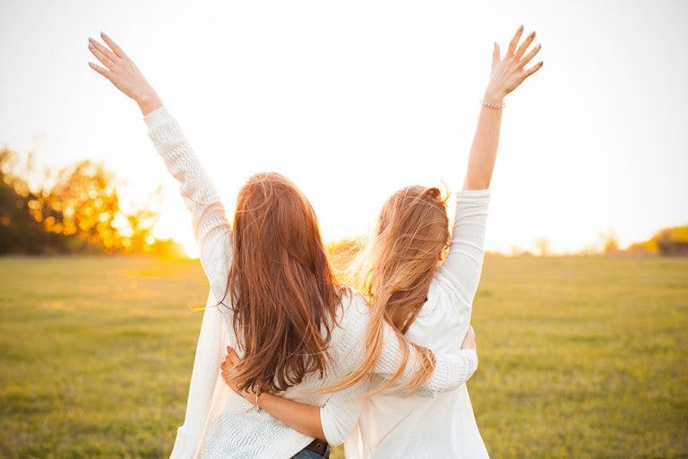 Two female best friends embracing each other’s back and lift their free hands into the air
