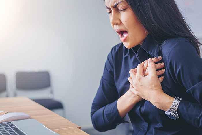 Woman in workwear clutching her chest as she grimaces in pain while sitting at her work desk