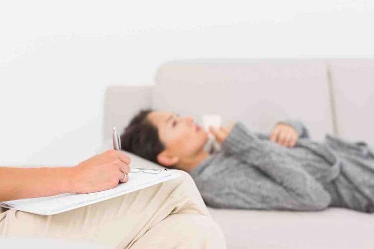 A woman lies down on a couch and holds a cup while a psychiatrist writes down information on a notepad