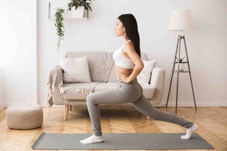 An Asian woman in sports attire does bodyweight lunges on a grey yoga mat in her living room