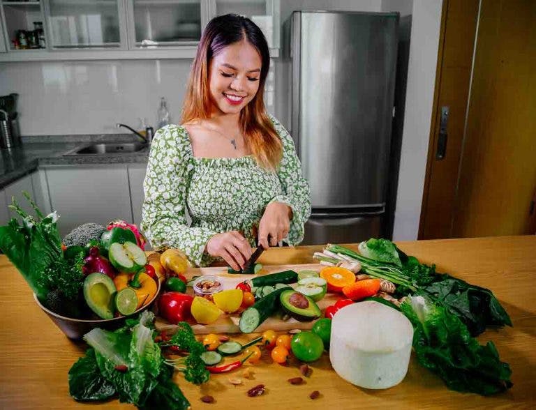 An Asian woman cutting vegetables on a kitchen counter filled with fruits and vegetables