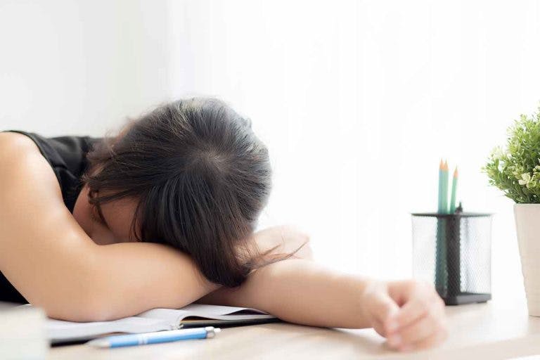 A young woman is asleep on her desk, and her head is resting on her arms on top of a book