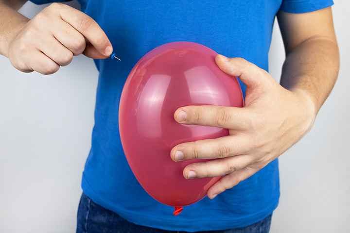 Man in blue shirt holds a red balloon in left hand and a thumbtack in his right hand