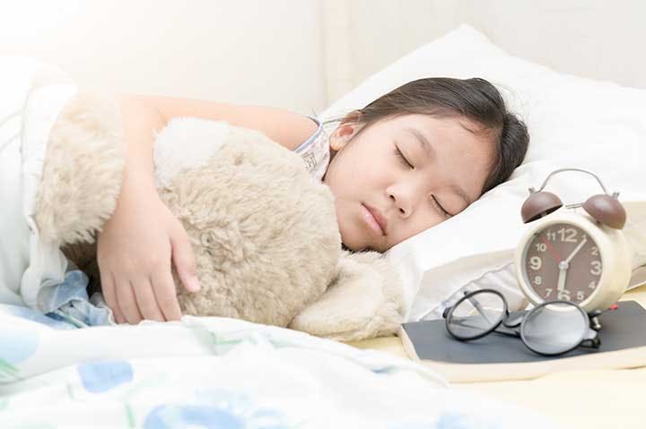 Little girl sleeping soundly with her teddy bear and an alarm clock next to her