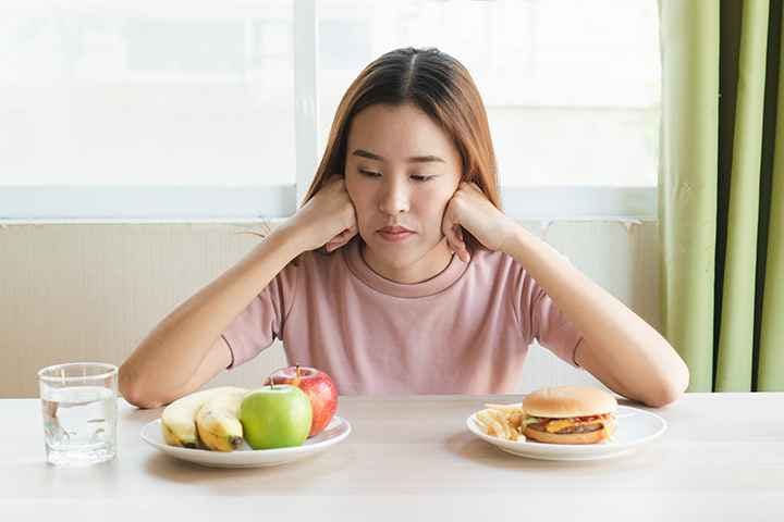 A woman looks seemingly indecisive about a plate of burgers and a plate of fruits on the table in front of her