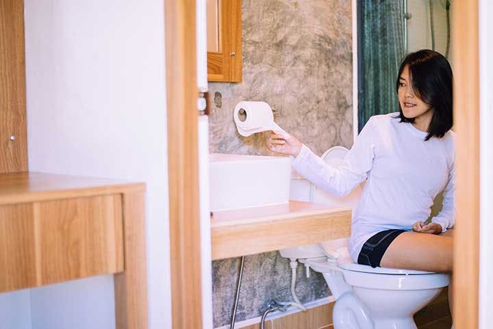 A woman sitting on the toilet and reaching out to a roll of toilet paper behind her