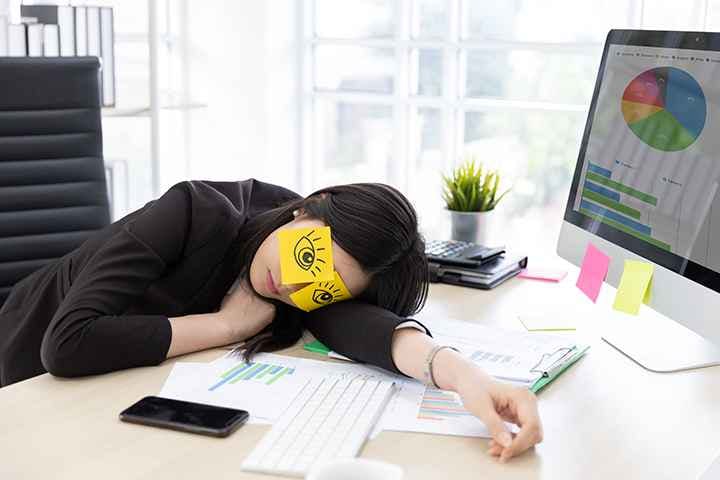 A woman sleeping on her desk at work with drawings of opened eyes placed over her real eyes