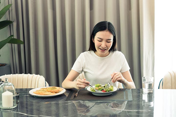 A young woman at a dining table, smiling as she takes a scoop of her salad