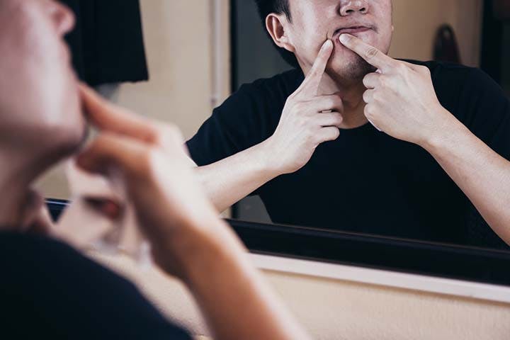 Young man squeezing acne on his chin while looking in the mirror 