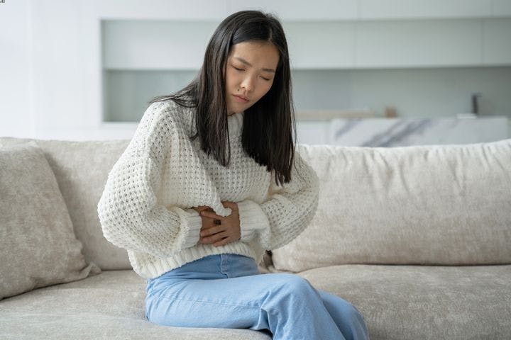 An Asian woman feeling abdominal pain while sitting on a sofa.