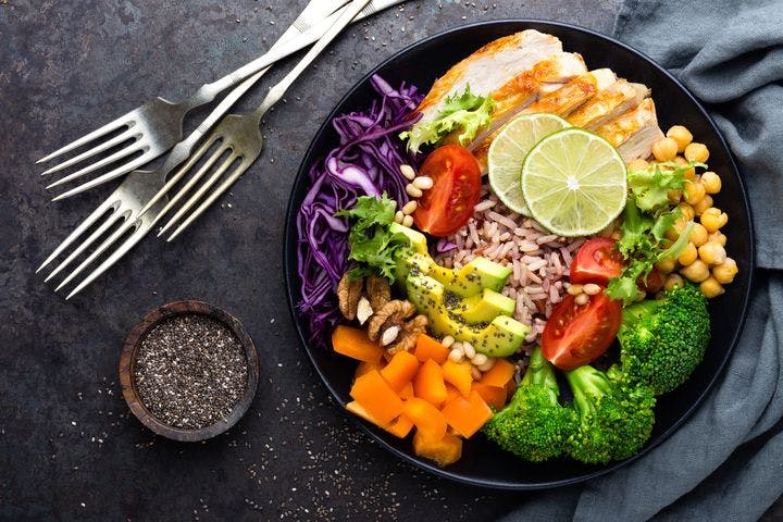 A plate full of healthy food. Brown rice topped with broccoli, carrots, purple cabbage, walnuts, pinenuts, chickpeas, avocado, tomato, lettuce and grilled chicken breast