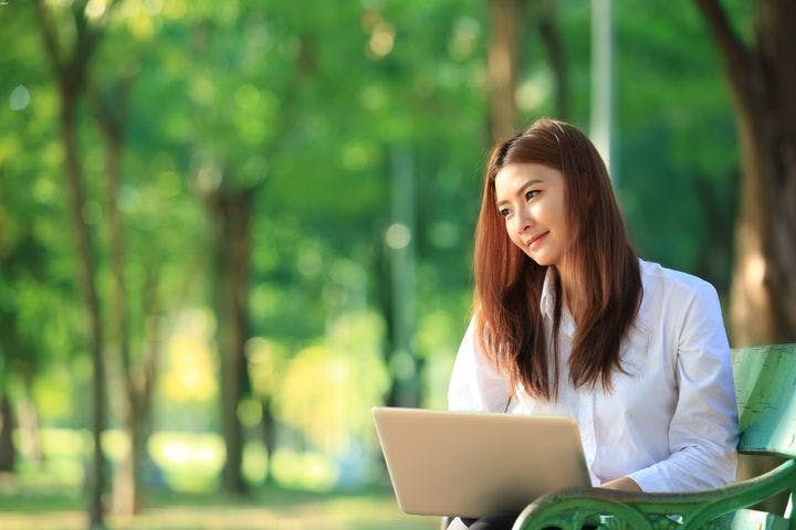 A young Asian lady is sitting on a bench and working on her laptop in a park