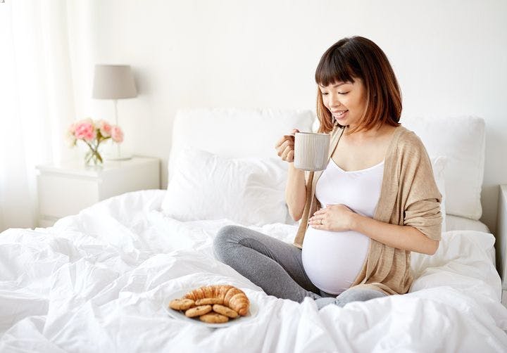 A pregnant woman sitting cross-legged on her bed while holding a cup of tea; a plate of biscuits in front of her