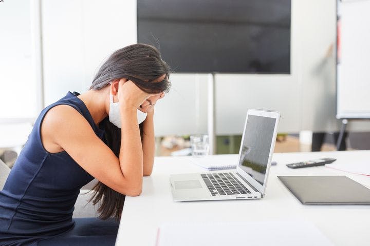 Woman wearing a white facial mask while holding her head in her hands as she sits at a table in front of her laptop.