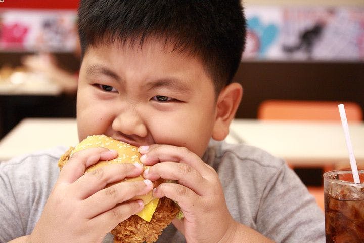 An Asian boy is eating a fried chicken burger with a glass of soda